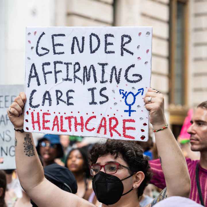 A shirtless demonstrator (wearing glasses and a black surgical mask) holds up a sign that reads" GENDER AFFIRMING CARE IS HEALTHCARE".